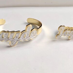 Puede incluir: Conjunto de pulsera y anillo de oro con el nombre "Jayde" escrito en cursiva y cubierto de purpurina blanca.