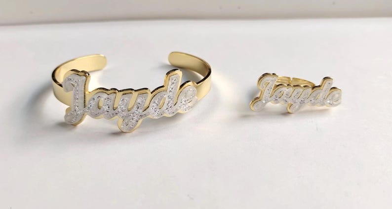 Puede incluir: Conjunto de pulsera y anillo de oro con el nombre "Jayde" escrito en cursiva y cubierto de purpurina blanca.