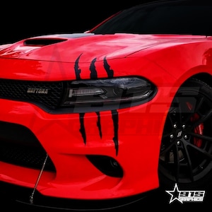 Può includere: Una Dodge Charger Daytona rossa con grafiche nere a forma di artigli sul lato del cofano. L'auto ha ruote nere e una griglia nera.