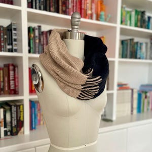 May include: A two-tone infinity scarf displayed on a mannequin. The scarf is beige and navy blue, with a knitted texture and fringed edges on the navy section. The background features a white bookshelf filled with books.