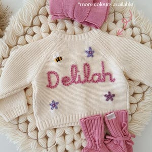 K&ouml;nnte beinhalten: Cremefarbener Baby-Pullover mit dem Namen &bdquo;Delilah&ldquo; in dunklem Rosa gestickt, verziert mit einer Biene und Sternen. Inklusive passender rosa Babyschuhe und einer Schleife. Der Text lautet &bdquo;Passendes Zubeh&ouml;r in anderen Angeboten erh&auml;ltlich&ldquo; und &bdquo;weitere Farben verf&uuml;gbar&ldquo;.