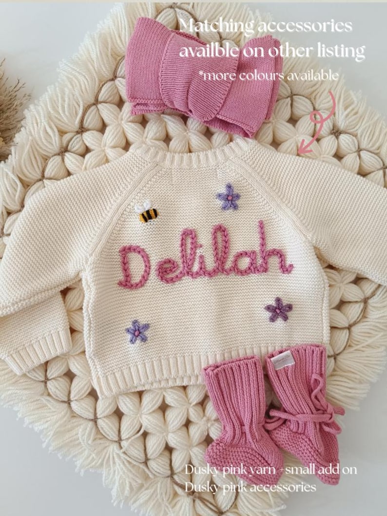 K&ouml;nnte beinhalten: Cremefarbener Baby-Pullover mit dem Namen &bdquo;Delilah&ldquo; in dunklem Rosa gestickt, verziert mit einer Biene und Sternen. Inklusive passender rosa Babyschuhe und einer Schleife. Der Text lautet &bdquo;Passendes Zubeh&ouml;r in anderen Angeboten erh&auml;ltlich&ldquo; und &bdquo;weitere Farben verf&uuml;gbar&ldquo;.