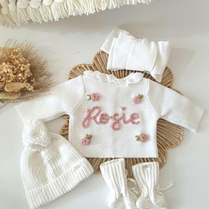 May include: A white knitted baby outfit set, including a sweater with the name "Rosie" embroidered in pink, a matching hat with a pom-pom, a headband, and booties. The sweater is adorned with small pink flower appliqu&eacute;s. The set is displayed on a woven mat.