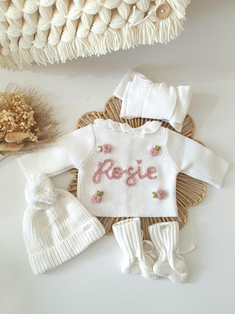 May include: A white knitted baby outfit set, including a sweater with the name "Rosie" embroidered in pink, a matching hat with a pom-pom, a headband, and booties. The sweater is adorned with small pink flower appliqu&eacute;s. The set is displayed on a woven mat.
