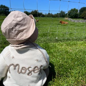 May include: A baby wearing a beige sun hat and a cream-colored sweater with the name "Moser" embroidered on the back in brown thread. The sweater has small star details. The baby is sitting in a grassy field with a fence and trees in the background.