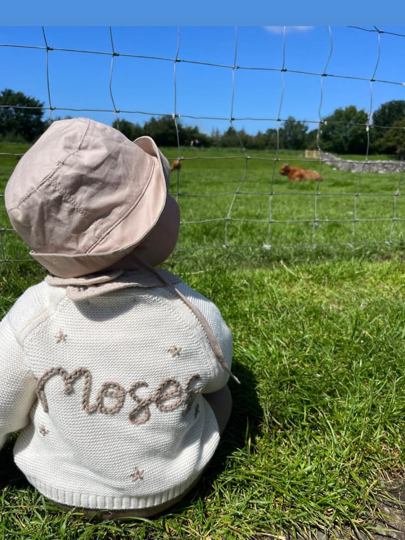 May include: A baby wearing a beige sun hat and a cream-colored sweater with the name "Moser" embroidered on the back in brown thread. The sweater has small star details. The baby is sitting in a grassy field with a fence and trees in the background.
