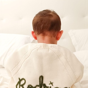 K&ouml;nnte beinhalten: Cremefarbener Strickpullover mit dem Namen "Rodrigo" in Olivgr&uuml;n gestickt. Der Pullover wird von hinten gesehen, wobei der Kopf und die Ohren des Kindes sichtbar sind. Der Pullover liegt auf einem wei&szlig;en Bett.