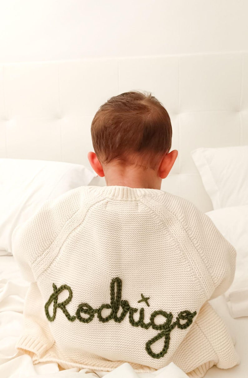 K&ouml;nnte beinhalten: Cremefarbener Strickpullover mit dem Namen "Rodrigo" in Olivgr&uuml;n gestickt. Der Pullover wird von hinten gesehen, wobei der Kopf und die Ohren des Kindes sichtbar sind. Der Pullover liegt auf einem wei&szlig;en Bett.