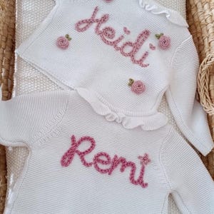 May include: Two white knit sweaters with long sleeves and ruffled collars. The sweaters are embroidered with names in pink yarn, one reading "Heidi" and the other "Remi." Each sweater is adorned with small pink rose appliques. The text at the bottom reads "Heidi" Pale Rose Yarn and "Remi" Dusky Pink Yarn.