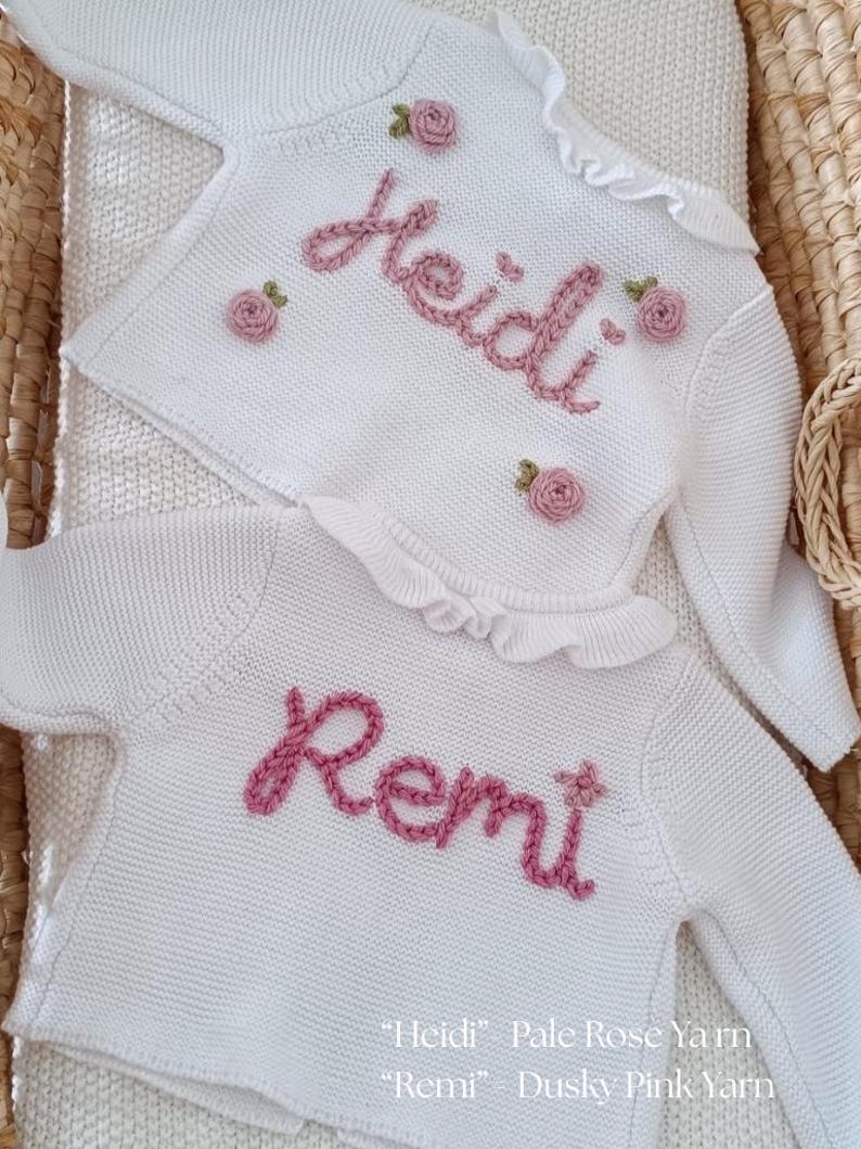 May include: Two white knit sweaters with long sleeves and ruffled collars. The sweaters are embroidered with names in pink yarn, one reading "Heidi" and the other "Remi." Each sweater is adorned with small pink rose appliques. The text at the bottom reads "Heidi" Pale Rose Yarn and "Remi" Dusky Pink Yarn.