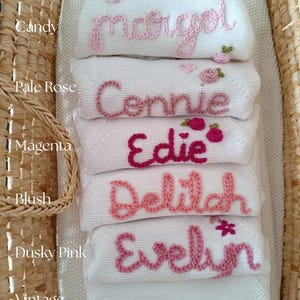 May include: A stack of white baby blankets embroidered with names in various shades of pink. The names include Margot, Connie, Edie, Delilah, Evelyn, and Cove. Each blanket has a different shade of pink embroidery.