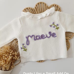 May include: A white knit sweater with a ruffled collar and long sleeves. The name "maeve" is embroidered in purple script across the front, with small floral accents. The text "Dusty Lilac + Small Add On" is at the bottom.