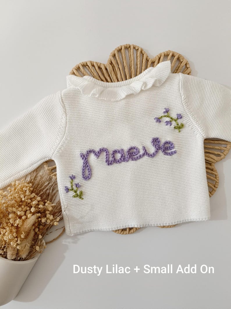May include: A white knit sweater with a ruffled collar and long sleeves. The name "maeve" is embroidered in purple script across the front, with small floral accents. The text "Dusty Lilac + Small Add On" is at the bottom.