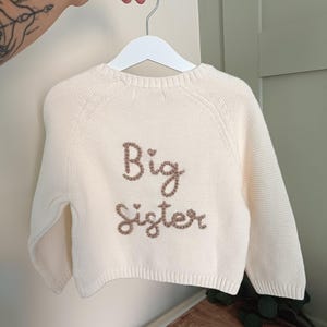 May include: Cream-colored knit sweater with the words "Big Sister" embroidered in brown thread. The sweater is hanging on a white hanger. The background is a light-colored wall.