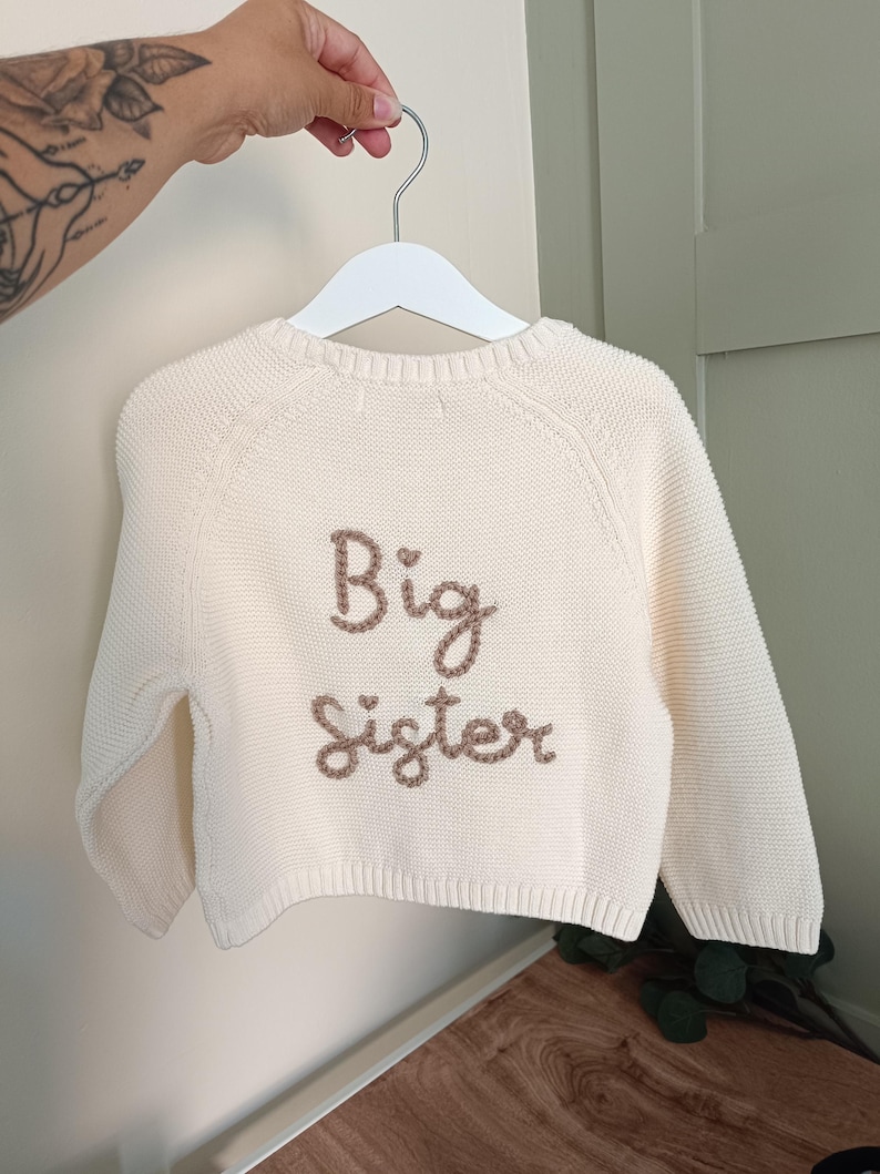 May include: Cream-colored knit sweater with the words "Big Sister" embroidered in brown thread. The sweater is hanging on a white hanger. The background is a light-colored wall.
