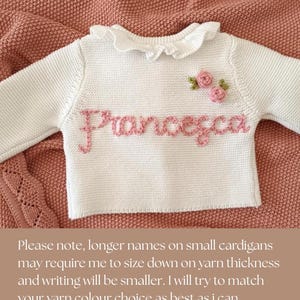May include: White baby sweater with the name "Francesca" embroidered in pink. The sweater has a ruffled collar and two small pink rose appliqu&eacute;s. The sweater is displayed on a pink knitted blanket.