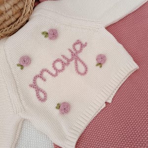 K&ouml;nnte beinhalten: Cremefarbene Baby-Strickjacke mit dem Namen "Maya" in rosa Schrift gestickt. Die Jacke ist mit kleinen, gestickten rosa Rosen und gr&uuml;nen Bl&auml;ttern verziert. Das Kleidungsst&uuml;ck wird auf einer rosa Decke pr&auml;sentiert.