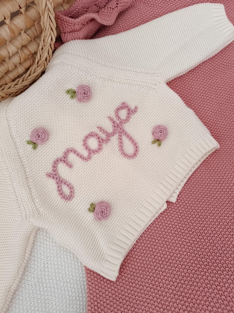 K&ouml;nnte beinhalten: Cremefarbene Baby-Strickjacke mit dem Namen "Maya" in rosa Schrift gestickt. Die Jacke ist mit kleinen, gestickten rosa Rosen und gr&uuml;nen Bl&auml;ttern verziert. Das Kleidungsst&uuml;ck wird auf einer rosa Decke pr&auml;sentiert.