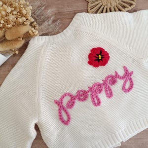 K&ouml;nnte beinhalten: Cremefarbener Strickpullover mit dem Wort "Poppy" aus rosa Garn gestickt. Auf der Brust ist eine rot-schwarze Blume gestickt. Der Pullover hat einen gerippten Saum und B&uuml;ndchen.