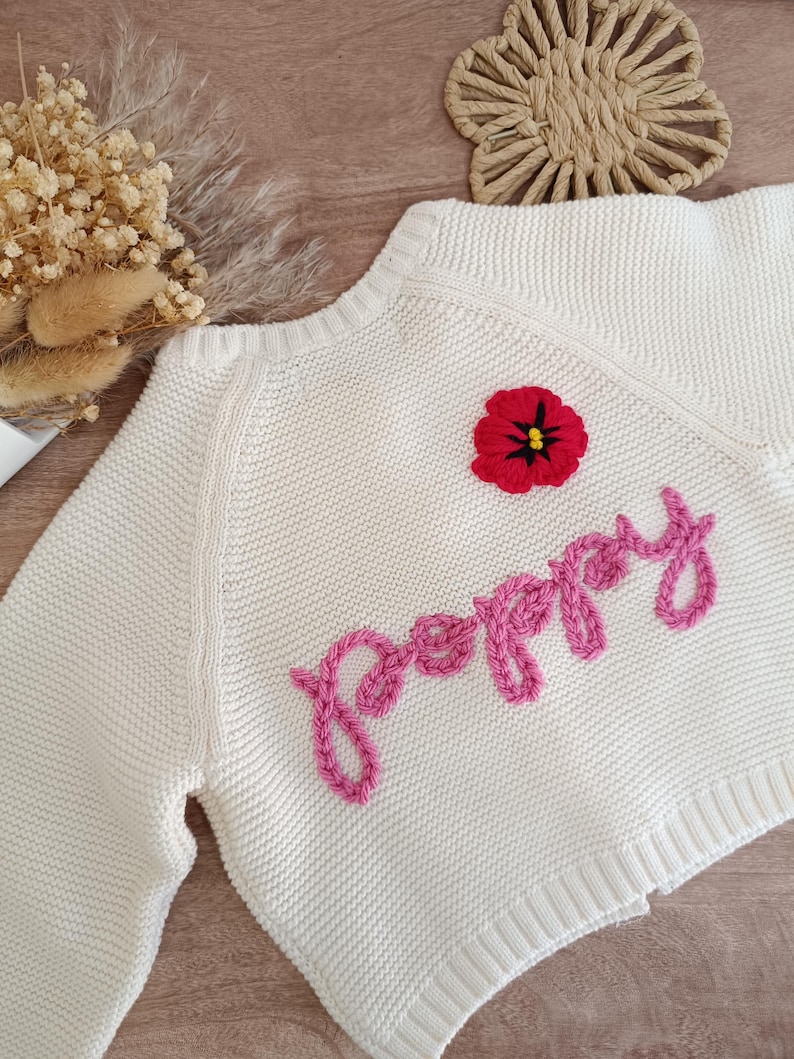 K&ouml;nnte beinhalten: Cremefarbener Strickpullover mit dem Wort "Poppy" aus rosa Garn gestickt. Auf der Brust ist eine rot-schwarze Blume gestickt. Der Pullover hat einen gerippten Saum und B&uuml;ndchen.