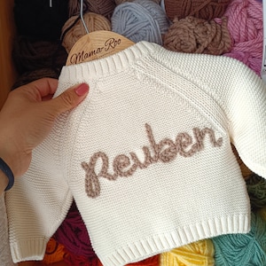 K&ouml;nnte beinhalten: Cremefarbener Baby-Pullover mit dem Namen "Reuben" in brauner Schrift gestickt. Der Pullover wird auf einem Holzb&uuml;gel mit dem Text "Mama Roo" pr&auml;sentiert. Der Hintergrund zeigt verschiedene Farben von Garn.