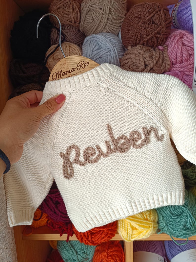 K&ouml;nnte beinhalten: Cremefarbener Baby-Pullover mit dem Namen "Reuben" in brauner Schrift gestickt. Der Pullover wird auf einem Holzb&uuml;gel mit dem Text "Mama Roo" pr&auml;sentiert. Der Hintergrund zeigt verschiedene Farben von Garn.