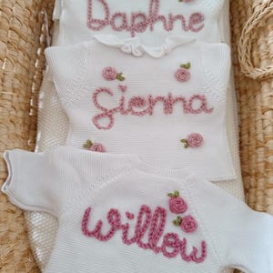 May include: Three white knit baby sweaters with embroidered names and pink rose accents. The names "Daphne", "Sienna", and "Willow" are stitched in a cursive font. The sweaters are displayed in a woven basket.