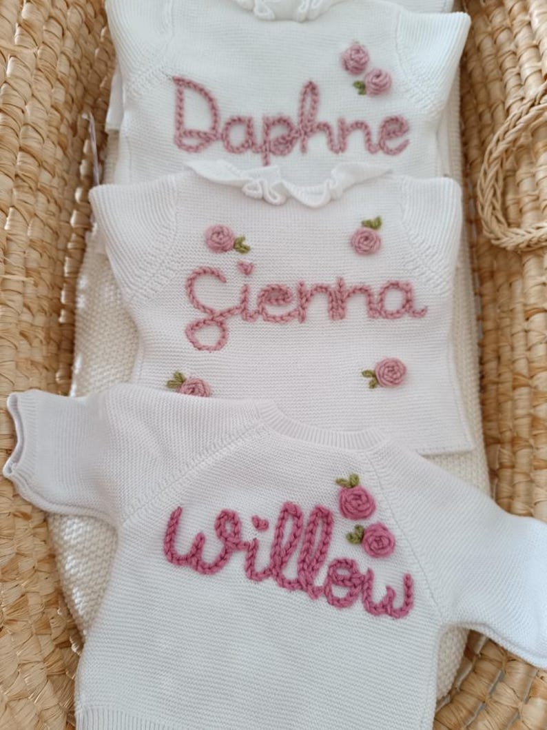May include: Three white knit baby sweaters with embroidered names and pink rose accents. The names "Daphne", "Sienna", and "Willow" are stitched in a cursive font. The sweaters are displayed in a woven basket.