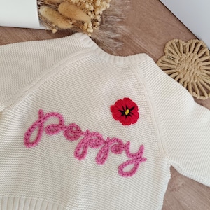 May include: Cream-colored knitted baby cardigan with the name "Poppy" embroidered in pink cursive. A red poppy flower with a black center is embroidered above the name. The cardigan has long sleeves and a ribbed hem.
