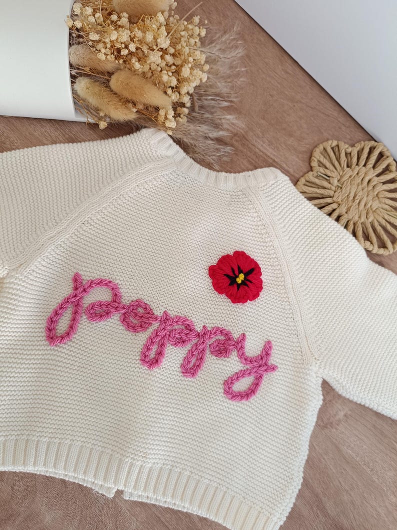 May include: Cream-colored knitted baby cardigan with the name "Poppy" embroidered in pink cursive. A red poppy flower with a black center is embroidered above the name. The cardigan has long sleeves and a ribbed hem.