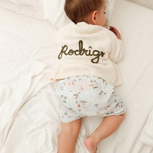 May include: A sleeping baby wearing a cream-colored cardigan with the name "Rodrigo" embroidered in green. The baby is also wearing light blue shorts with a safari animal print. The baby is lying on a white bed.