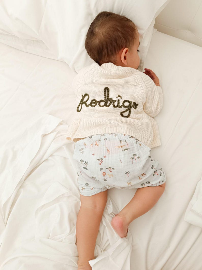 May include: A sleeping baby wearing a cream-colored cardigan with the name "Rodrigo" embroidered in green. The baby is also wearing light blue shorts with a safari animal print. The baby is lying on a white bed.
