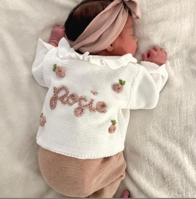 May include: A newborn baby wearing a white knit sweater with the name "Rosie" embroidered in pink. The sweater has pink rose appliqu&eacute;s and a ruffled collar. The baby is also wearing pink pants and a matching headband.