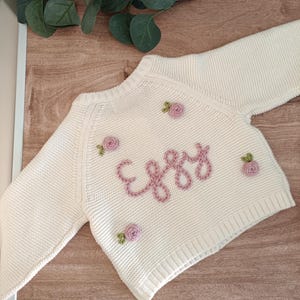 K&ouml;nnte beinhalten: Cremefarbener Baby-Pullover mit dem Namen "Eggy" in rosa Schrift gestickt. Der Pullover ist mit kleinen, gestickten rosa Rosen verziert. Das lang&auml;rmlige Strickwarenst&uuml;ck wird auf einer Holzoberfl&auml;che pr&auml;sentiert.
