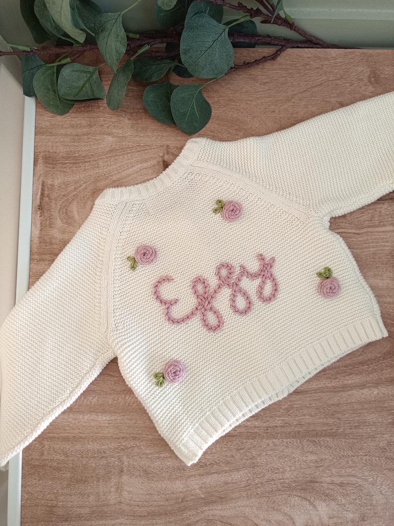 K&ouml;nnte beinhalten: Cremefarbener Baby-Pullover mit dem Namen "Eggy" in rosa Schrift gestickt. Der Pullover ist mit kleinen, gestickten rosa Rosen verziert. Das lang&auml;rmlige Strickwarenst&uuml;ck wird auf einer Holzoberfl&auml;che pr&auml;sentiert.