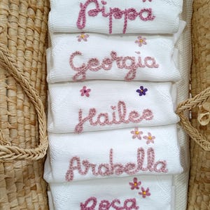 May include: White baby blankets embroidered with names in pink thread: Pippa, Georgia, Hailee, Arabella, and Rosa. Each blanket has small flower accents. The blankets are stacked inside a woven basket.
