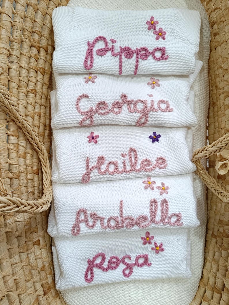 May include: White baby blankets embroidered with names in pink thread: Pippa, Georgia, Hailee, Arabella, and Rosa. Each blanket has small flower accents. The blankets are stacked inside a woven basket.