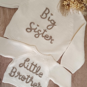 May include: Two cream-colored knit sweaters, one labeled "Big Sister" and the other "Little Brother" in brown embroidered lettering. The sweaters are displayed on a wooden surface, suggesting a baby or children's clothing theme.