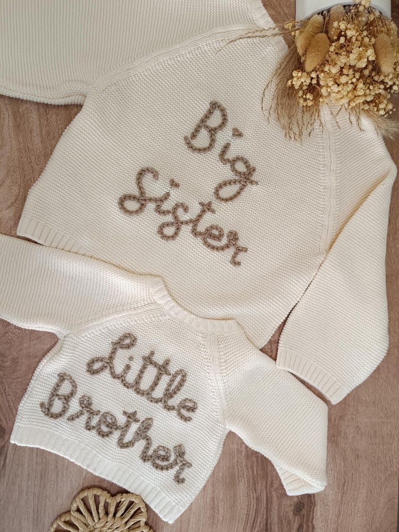 May include: Two cream-colored knit sweaters, one labeled "Big Sister" and the other "Little Brother" in brown embroidered lettering. The sweaters are displayed on a wooden surface, suggesting a baby or children's clothing theme.