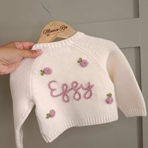 May include: Cream-colored baby sweater with the name "Eggy" embroidered in pink script. The sweater features small, embroidered pink roses. The sweater is displayed on a wooden hanger with the "Mama Roo" logo.