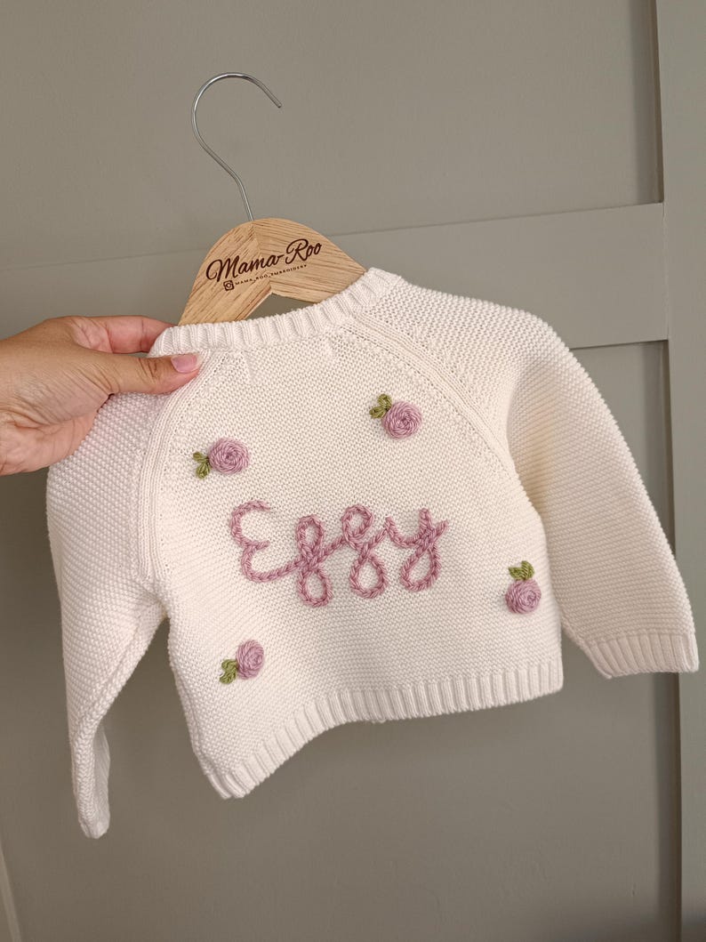 May include: Cream-colored baby sweater with the name "Eggy" embroidered in pink script. The sweater features small, embroidered pink roses. The sweater is displayed on a wooden hanger with the "Mama Roo" logo.