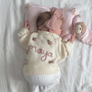 K&ouml;nnte beinhalten: Ein schlafendes Baby in einem cremefarbenen Pullover mit dem Namen "maya" in Rosa gestickt, zusammen mit rosa floralen Akzenten. Das Baby tr&auml;gt auch ein passendes rosa Schleifen-Stirnband und ruht auf einem rosa Kissen mit einem Teddyb&auml;ren. Das Baby tr&auml;gt eine wei&szlig;e Hose.