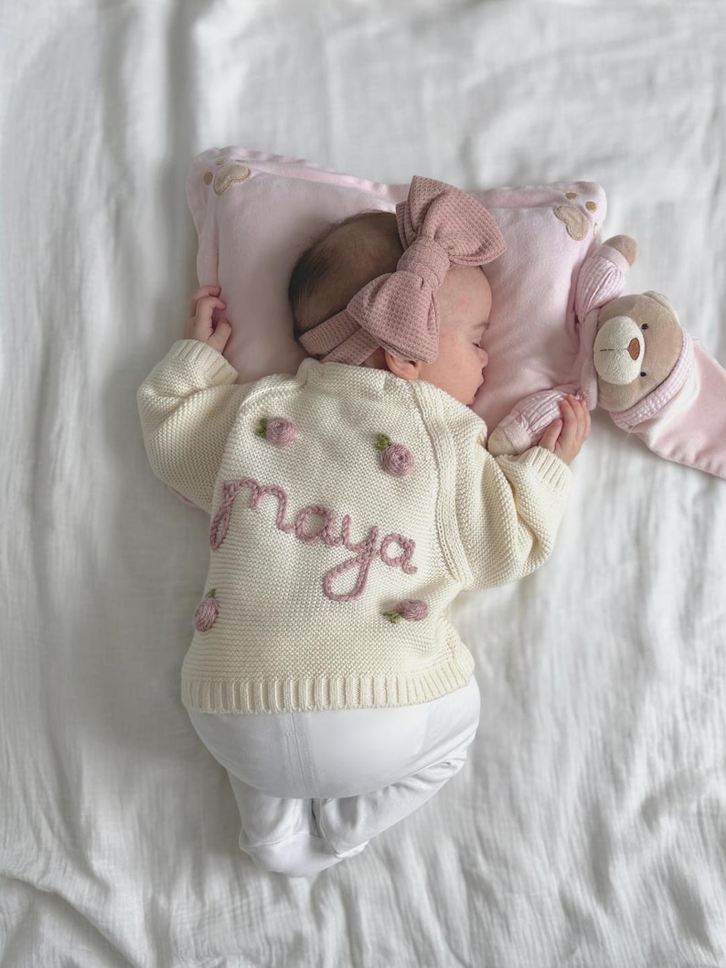 K&ouml;nnte beinhalten: Ein schlafendes Baby in einem cremefarbenen Pullover mit dem Namen "maya" in Rosa gestickt, zusammen mit rosa floralen Akzenten. Das Baby tr&auml;gt auch ein passendes rosa Schleifen-Stirnband und ruht auf einem rosa Kissen mit einem Teddyb&auml;ren. Das Baby tr&auml;gt eine wei&szlig;e Hose.