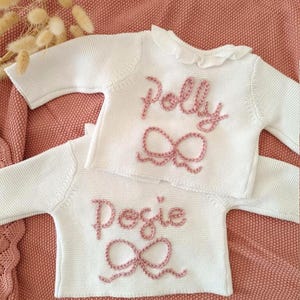 May include: Two white knitted baby sweaters with long sleeves, each embroidered with a name and a bow in pale pink yarn. The sweater on top says "Polly", and the bottom one says "Posie". The sweaters are on a pink knitted blanket. The text "Pale Rose Yarn" is at the bottom.