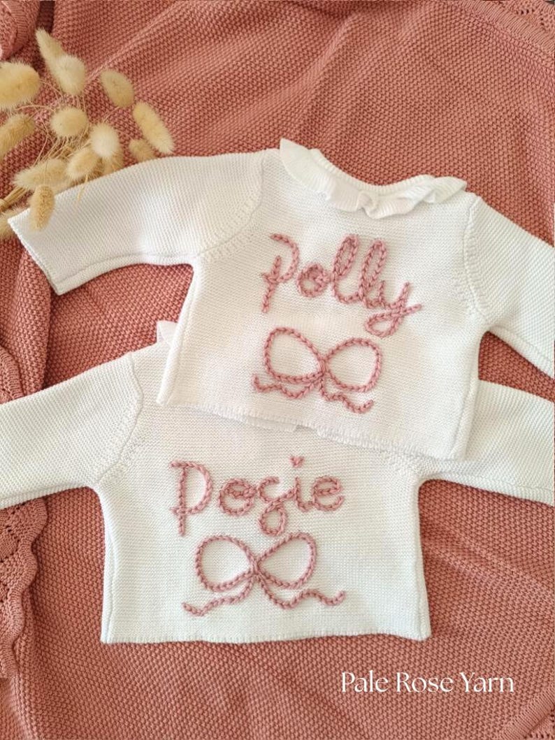 May include: Two white knitted baby sweaters with long sleeves, each embroidered with a name and a bow in pale pink yarn. The sweater on top says "Polly", and the bottom one says "Posie". The sweaters are on a pink knitted blanket. The text "Pale Rose Yarn" is at the bottom.