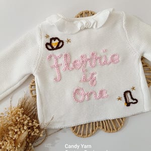 May include: White knit baby sweater with a ruffled collar. The sweater features embroidered text in pink that reads "Florrie is One", along with a cowboy hat and boot design. The text "Candy Yarn" is also visible.