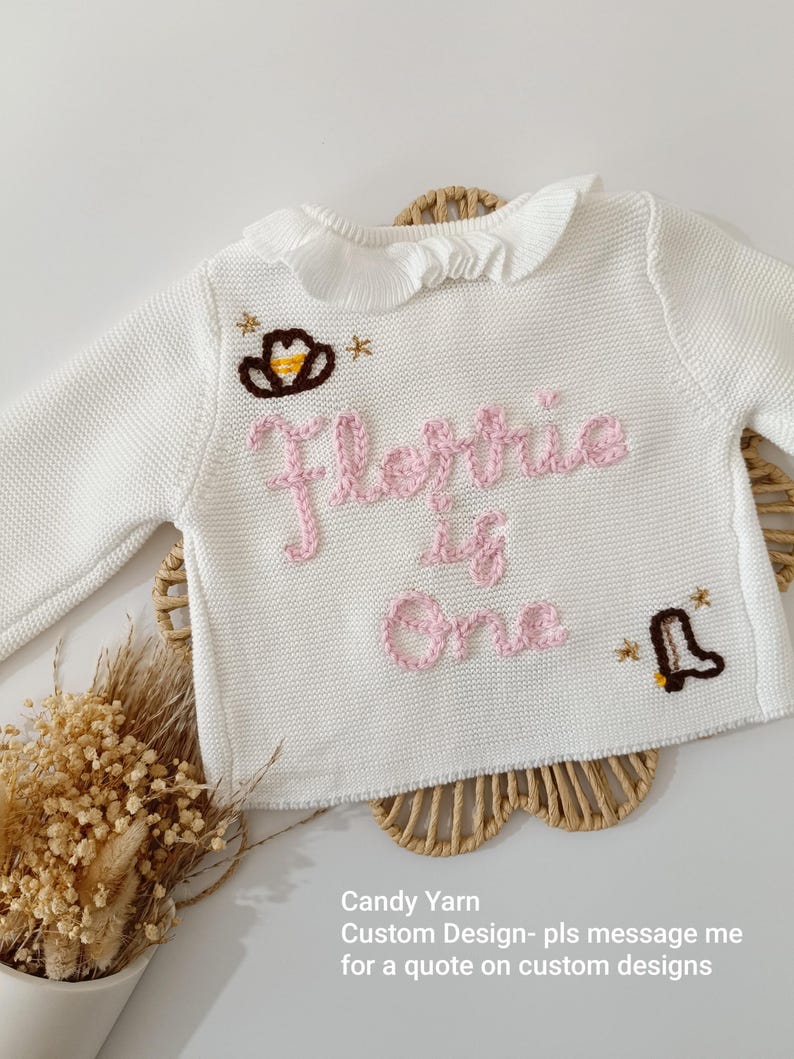 May include: White knit baby sweater with a ruffled collar. The sweater features embroidered text in pink that reads "Florrie is One", along with a cowboy hat and boot design. The text "Candy Yarn" is also visible.