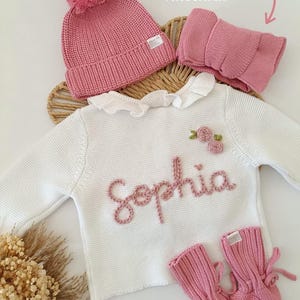 May include: A baby clothing set in pink and white. The set includes a knitted hat with a pom-pom, a neck warmer, a white sweater with the name "Sophia" embroidered in pink, and matching pink booties. The text in the image says "Matching accessories available on other listing *More colours".