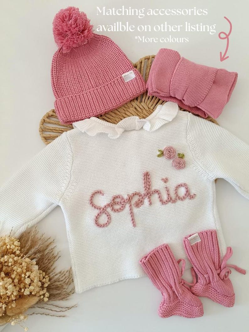 May include: A baby clothing set in pink and white. The set includes a knitted hat with a pom-pom, a neck warmer, a white sweater with the name "Sophia" embroidered in pink, and matching pink booties. The text in the image says "Matching accessories available on other listing *More colours".