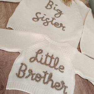 K&ouml;nnte beinhalten: Cremefarbene Strickpullover, einer mit der Aufschrift "Big Sister" und der andere mit "Little Brother" in brauner Stickerei. Die Pullover liegen auf einer hellen Holzoberfl&auml;che, mit einem kleinen Blumengesteck im Hintergrund.